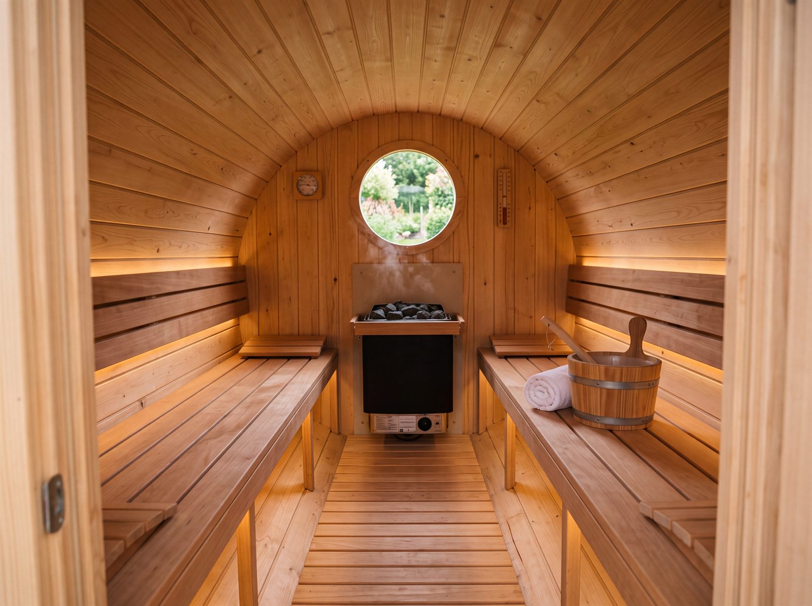 Innenraum einer Fasssauna mit Bänken aus hellem Holz, Saunaofen an der Stirnwand und rundem Fenster