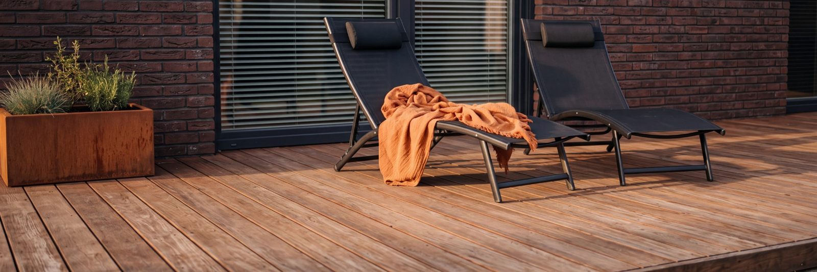 Holzterrasse mit zwei schwarzen Liegen und oranger Decke vor einer Backsteinwand