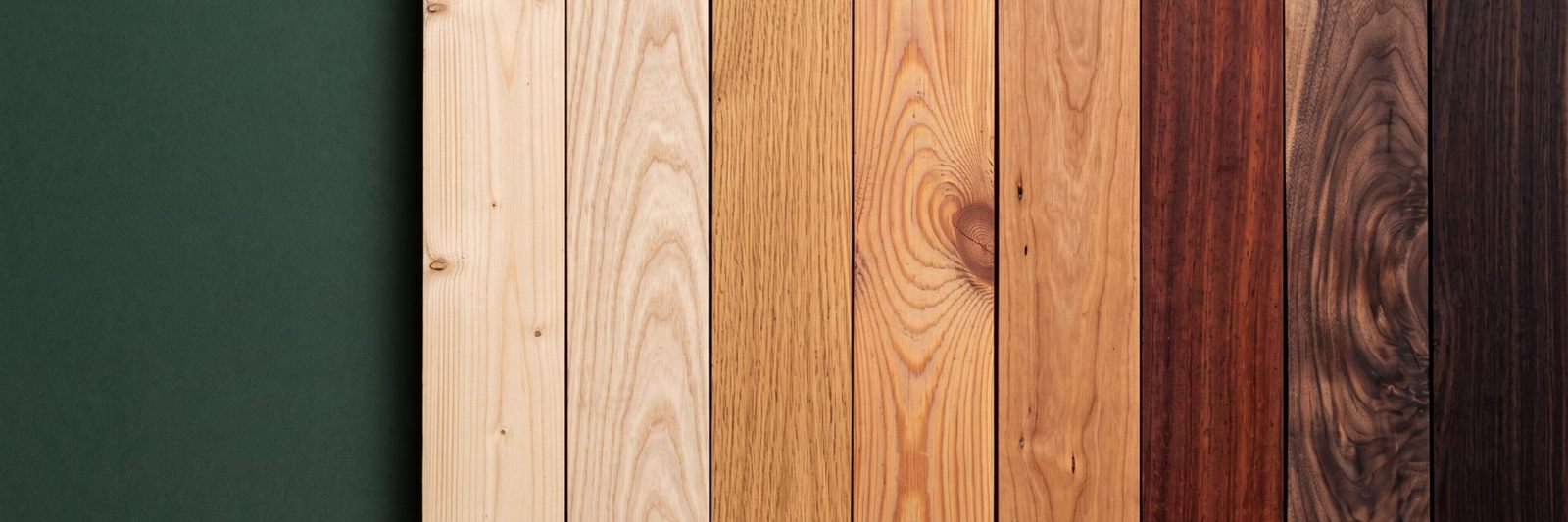 Holzbretter verschiedener Holzarten nebeneinander vor dunkelgrünem Hintergrund