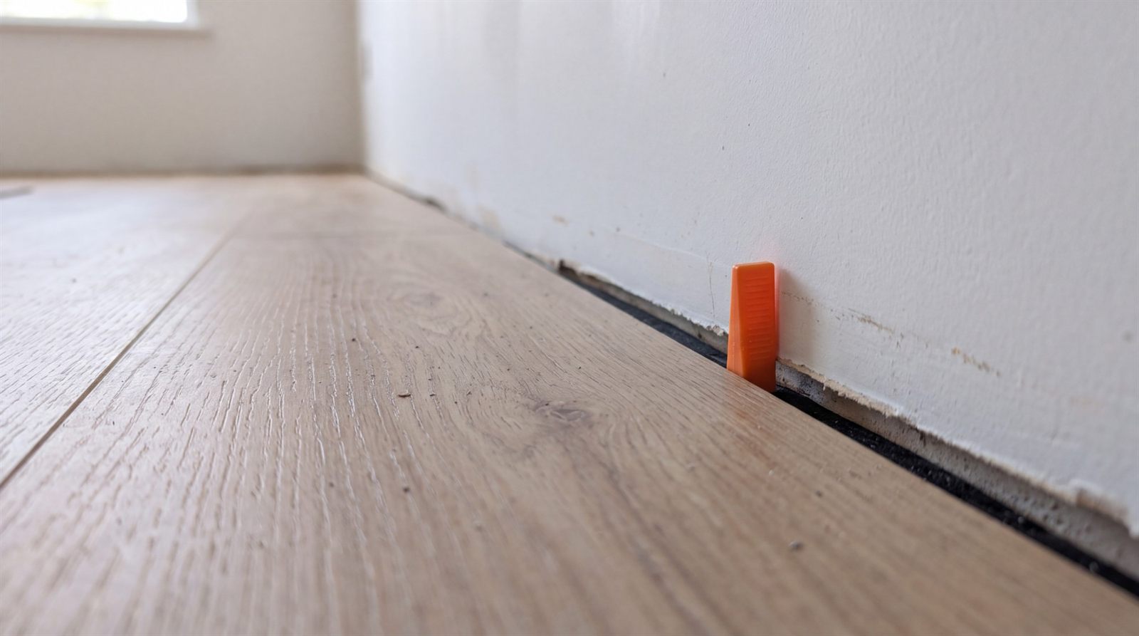Vinylplanken an einer Wand verlegt, ein Abstandskeil hält einen Spalt zwischen Planke und Wand frei